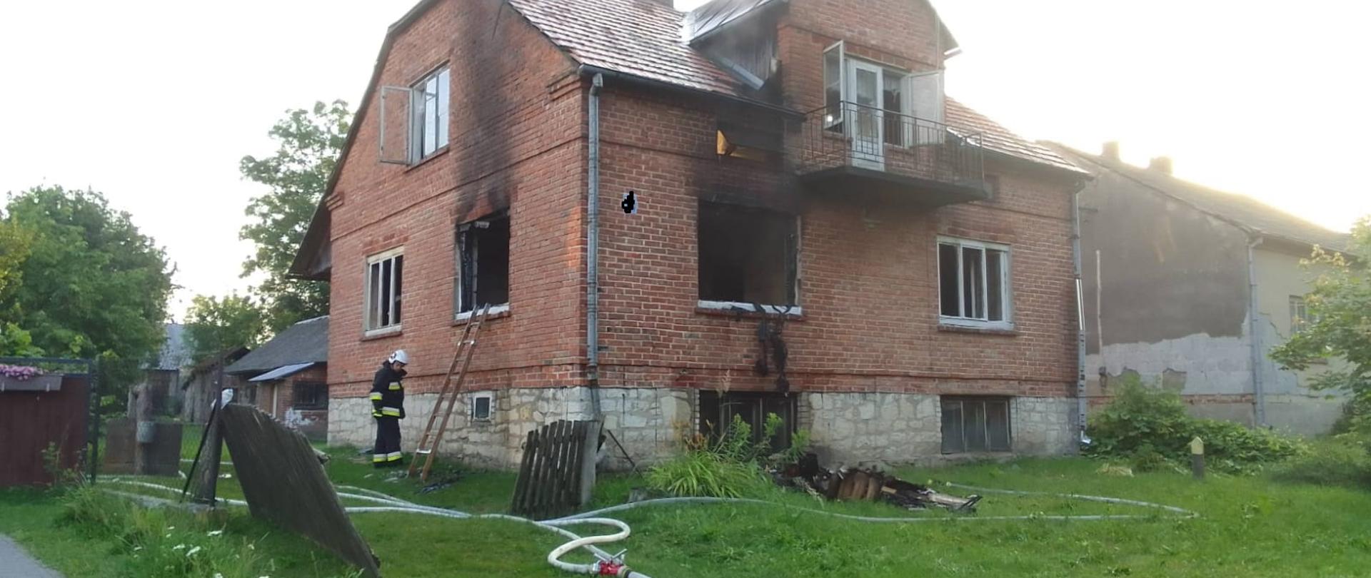 Zdjęcie przedstawia dom jednorodzinny wymurowany z cegły. Na budynku widoczne są ślady pożaru. Dom nie posiada dwóch okien.