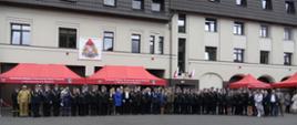 Grupa strażaków oraz zaproszonych gości stoi przed budynkiem Komendy Miejskiej Państwowej Straży Pożarnej w Siemianowicach Śląskich. Nad nimi widoczne są czerwone namioty z napisem komendy oraz baner z herbem straży pożarnej. W tle biały budynek z wieloma oknami i dwiema polskimi flagami.