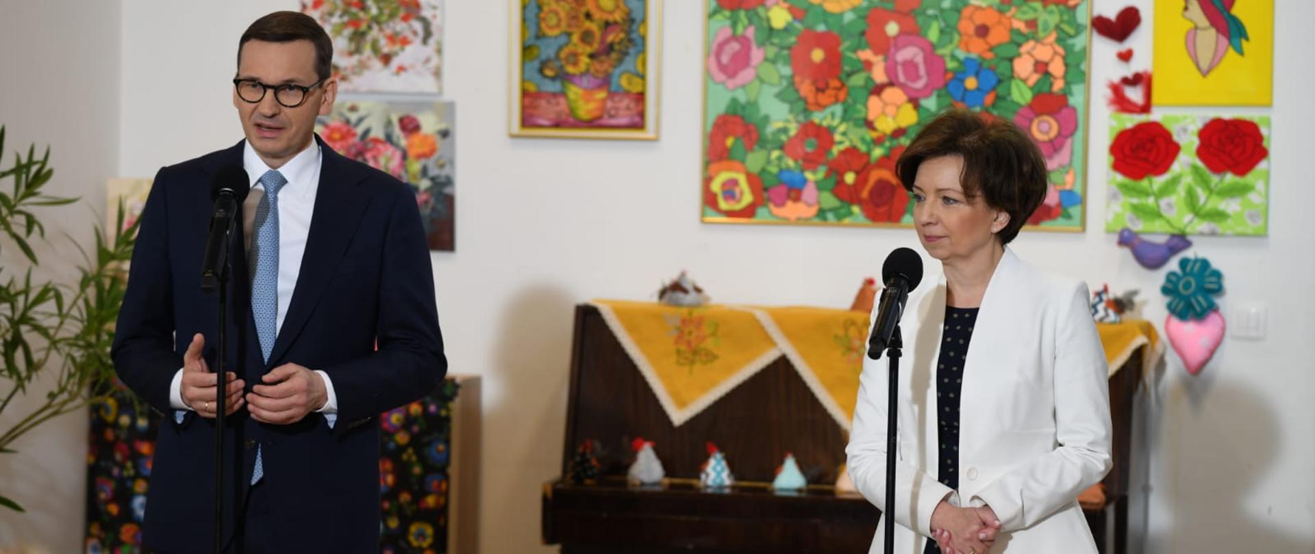Na zdjęciu premier Morawiecki i minister Maląg. Stoją przy mikrofonach. Z gestykulacji wynika, że przemawia właśnie premier Morawiecki. W tle na ścianie wiszą kolorowe obrazki. Widać także pianino, na którym zostaly umieszczone elementy świąteczne. 