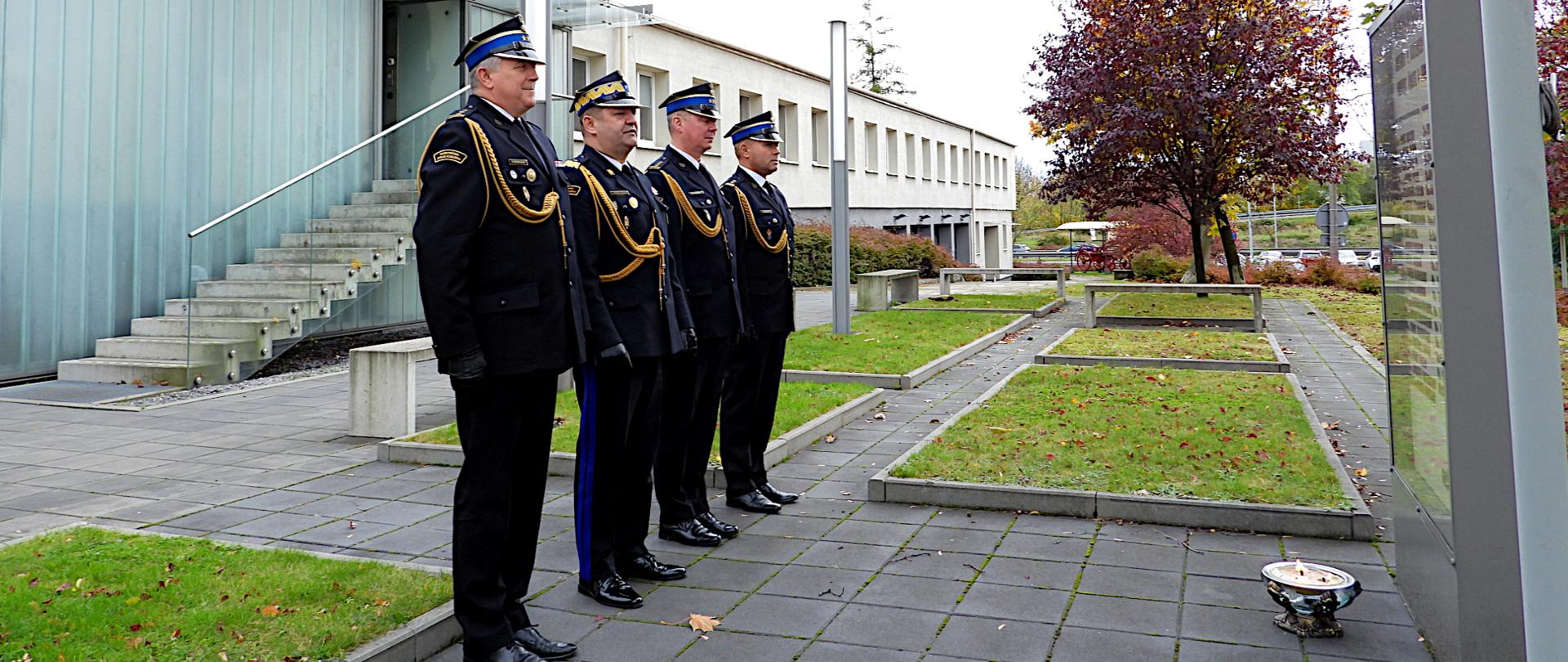 Śląski Komendant Wojewódzki PSP nadbryg. Jacek Kleszczewski w asyście swoich Zastępców st. bryg. Mirosława Synowca, bryg. Arkadiusza Krzemińskiego oraz st. bryg. Rafała Świerczka. Ubrani w mundury wyjściowe ze sznurami i rogatywki oddają hołd przed tablicą upamiętniającą poległych na służbie strażaków i druhów. Przed tablica, na ziemi zapalony znicz. Po lewej stronie parterowy budynek komendy wojewódzkiej i wejście główne do komendy. Wokół trawnik i chodnik.