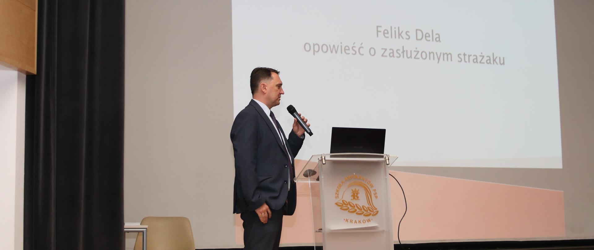 Prelegent – pracownik Centralnego Muzeum Pożarnictwa w Mysłowicach – Dariusz Falecki w czasie prezentacji. Ubrany w garnitur mężczyzna za pulpitem ustawionym na scenie szkolnej auli. 
