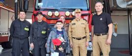 Na tle samochodu pożarniczego stoją w szeregu gość który odwiedził komendę powiatową psp w brzezinach czyli trzynastoletni letni Szymon wraz z mamą, obok stoi czterech strażaków wraz z komendantem powiatowym psp w brzezinach