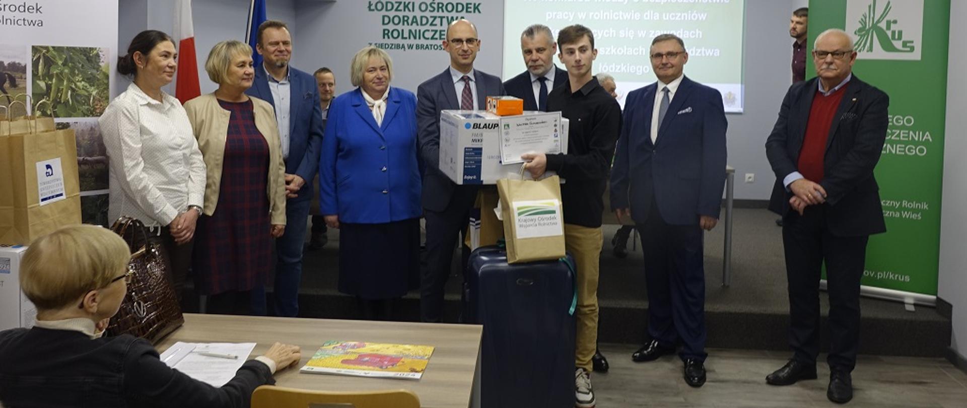 Laureat konkursu trzymający nagrody stoi przed przedstawicielami współorganizatorów.