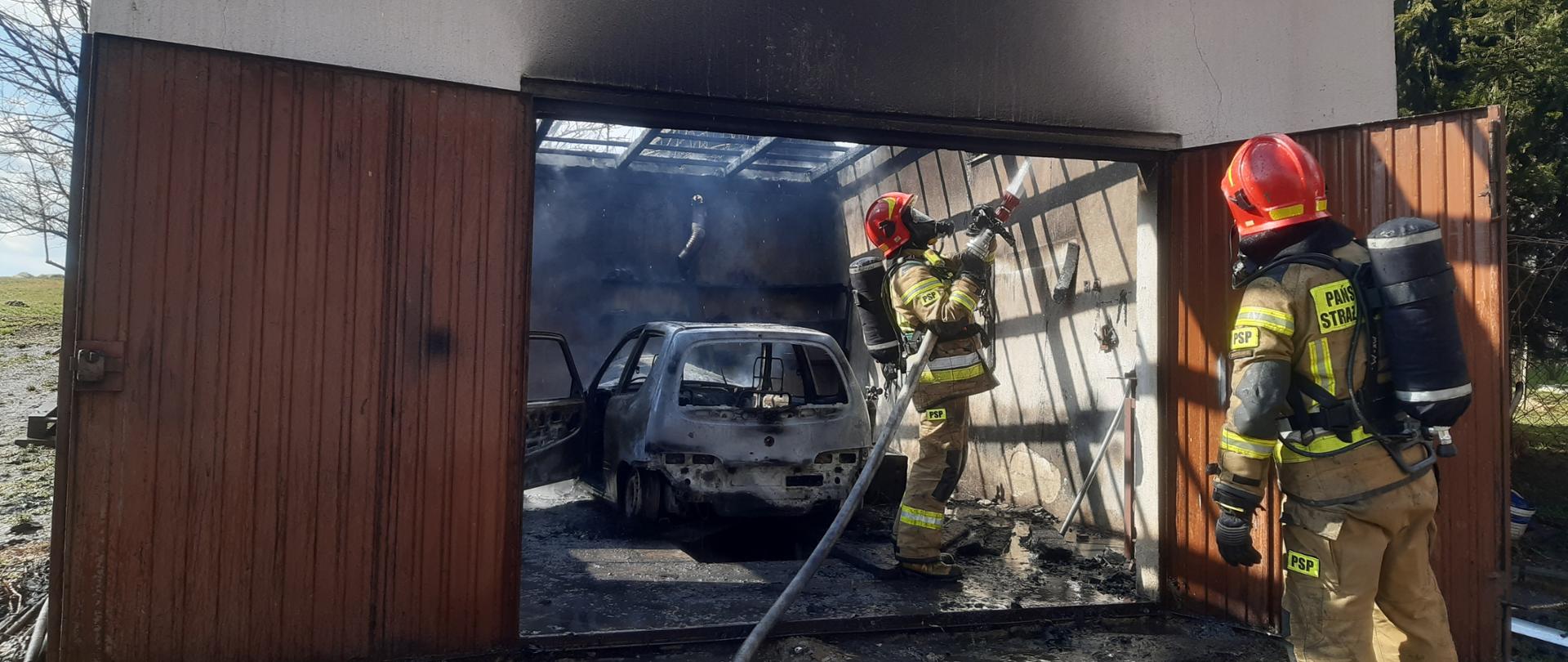 Zdjęcie przedstawia strażaków z linią gaśniczą na tle spalonego garażu oraz samochodu dogaszających pożar