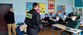 Zdjęcie przedstawia strażaków w trakcie zajęć z młodzieżą w klasie szkolnej. Na pierwszym planie po lewej stronie widoczny strażak w czarnym mundurze. Po prawej stronie widać grupę 16 dzieci siedzących w ławkach szkolnych. Na pierwszej ławce rozłożony jest fantom do ćwiczeń z resuscytacji krążeniowo-oddechowej.