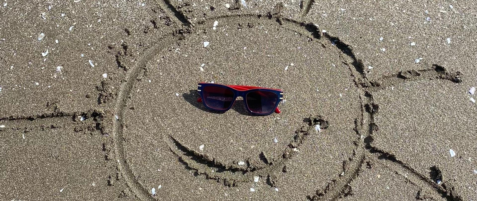 Grafika na której przedstawiono narysowane na brzegu plaży na piasku słoneczko z dołączonymi czarnymi okularami.