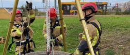 Na zdjęciu widać trzech strażaków mający na sobie piaskowe ubrania a na nich czerwono – czarne uprzęże do działań wysokościowych. Na rękach mają rękawice a na głowach czerwone kaski. Mężczyźni stoją przy studni z której ewakuują kolejną osobę. Dookoła studni rozstawione jest urządzenie o nazwie trójnóg ratowniczy do którego zamocowana jest lina.
W tle zdjęcia widzimy płot a za nim autobus jadący po drodze miejskiej.
Sytuacja dzieje się na trawniku.
