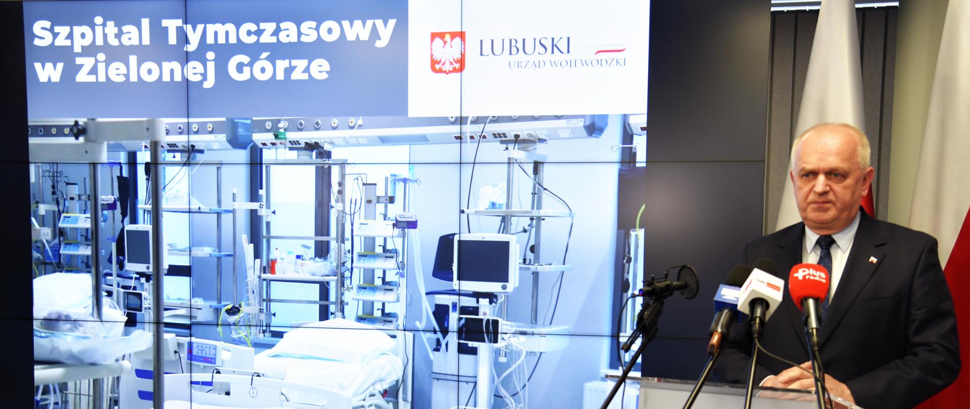 Wojewoda w czasie konferencji prasowej - szeroki widok na mównicę i ekran z prezentacją