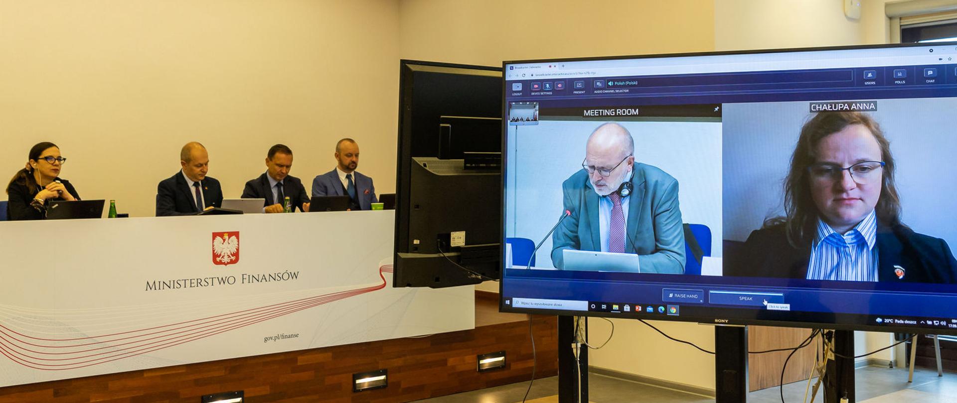 Osoby na ekranie podczas telekonferencji oraz uczestnicy przy stole prezydialnym.
