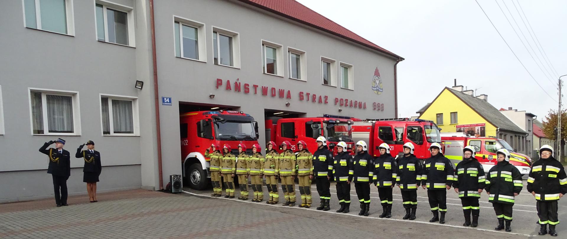 Zdjęcie wykonano przed budynkiem Komendy Powiatowej Państwowej Straży Pożarnej w Sępólnie Krajeńskim. Strażacy Państwowej i Ochotniczej Straży Pożarnej stoją w szeregu na baczność podczas wciągania flagi na maszt. Komendant Powiatowy, pani aspirant ubrani w mundury wyjściowe oraz dowódca zmiany ubrany w ubranie specjalne salutują. W tle samochody pożarnicze stoją przed garażami. 