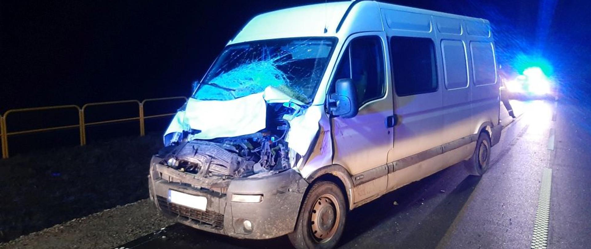 W nocy na drodze asfaltowej stoi biały bus. Pobita przednia szyba i zgnieciona część silnikowa. Z tyłu świecą światła innego samochodu.