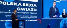 Minister Kulasek stoi za mównicą i mówi do mikrofonu, za nim niebieska ściana z napisem IGNIS - Polska sięga gwiazd.