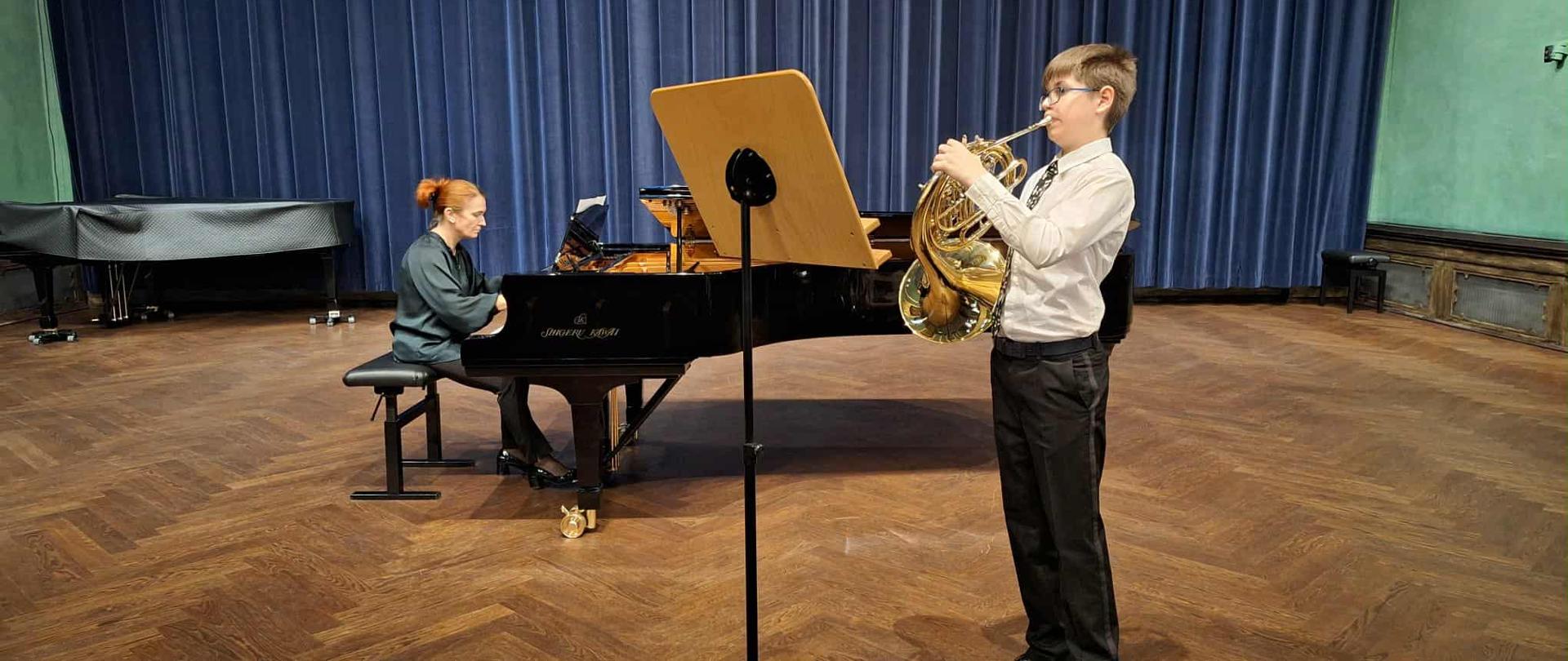 Uczeń gra na waltorni, akompaniuje nauczycielka przy fortepianie, w tle niebieska kotara, drewniana podłoga.