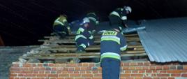 Na zdjęciu widać 5strażaków w ubraniach specjalnych. Strażacy zabezpieczają uszkodzony przez wiatr dach na budynku mieszkalnym. 4 strażaków znajduje się na dachu, a 1 na drabinie przystawionej do ściany budynku. Pora nocna. Teren akcji jest oświetlany.
