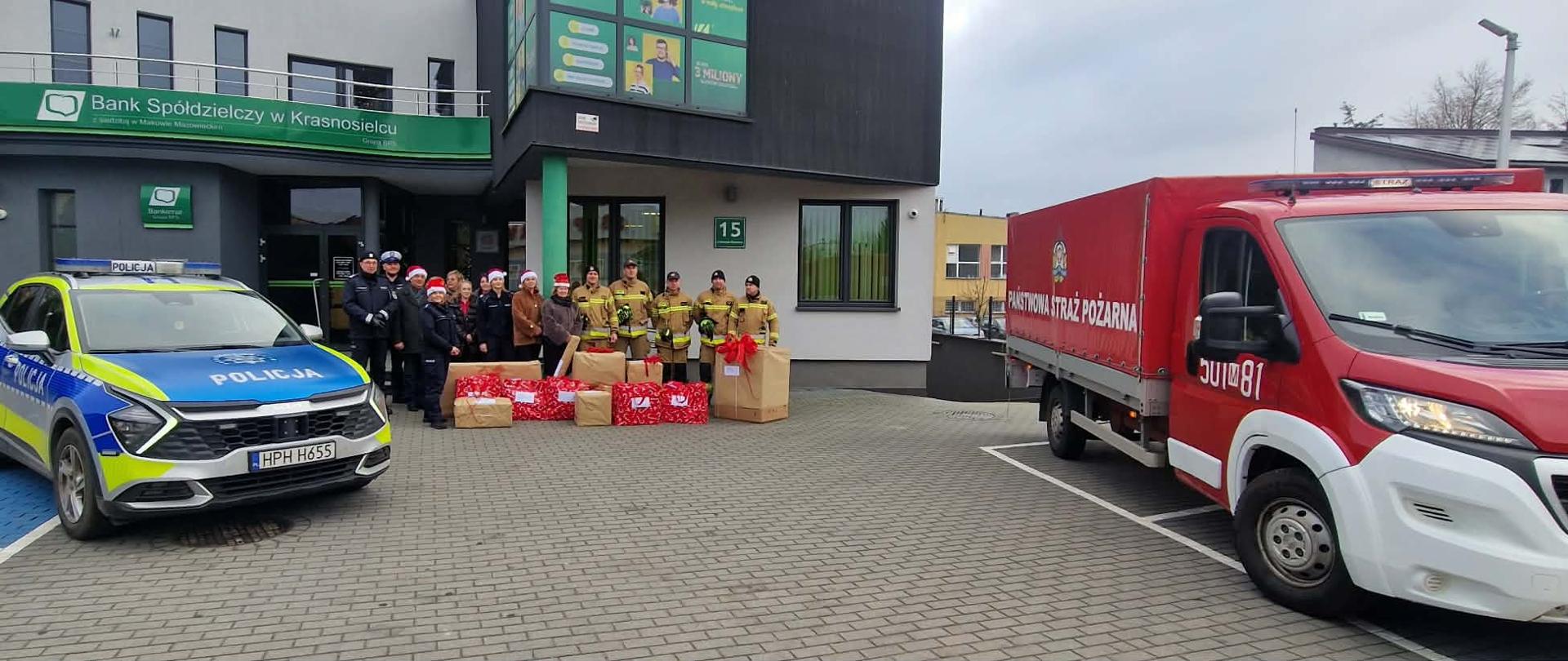 Wspólne zdjęcie strażaków, policjantów i pracowników banku z paczkami świątecznymi na tle budynku Komendy, pojazdy służbowe. 