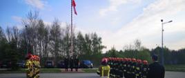 Na zdjęciu widoczni strażacy z Jednostki Ratowniczo-Gaśniczej nr 2 w Tarnowie podczas zbiórki. Strażacy w mundurach bojowych ustawieni w dwóch szpalerach, pośrodku widoczny maszt, poczet flagowy i podniesiona flaga państwowa. 