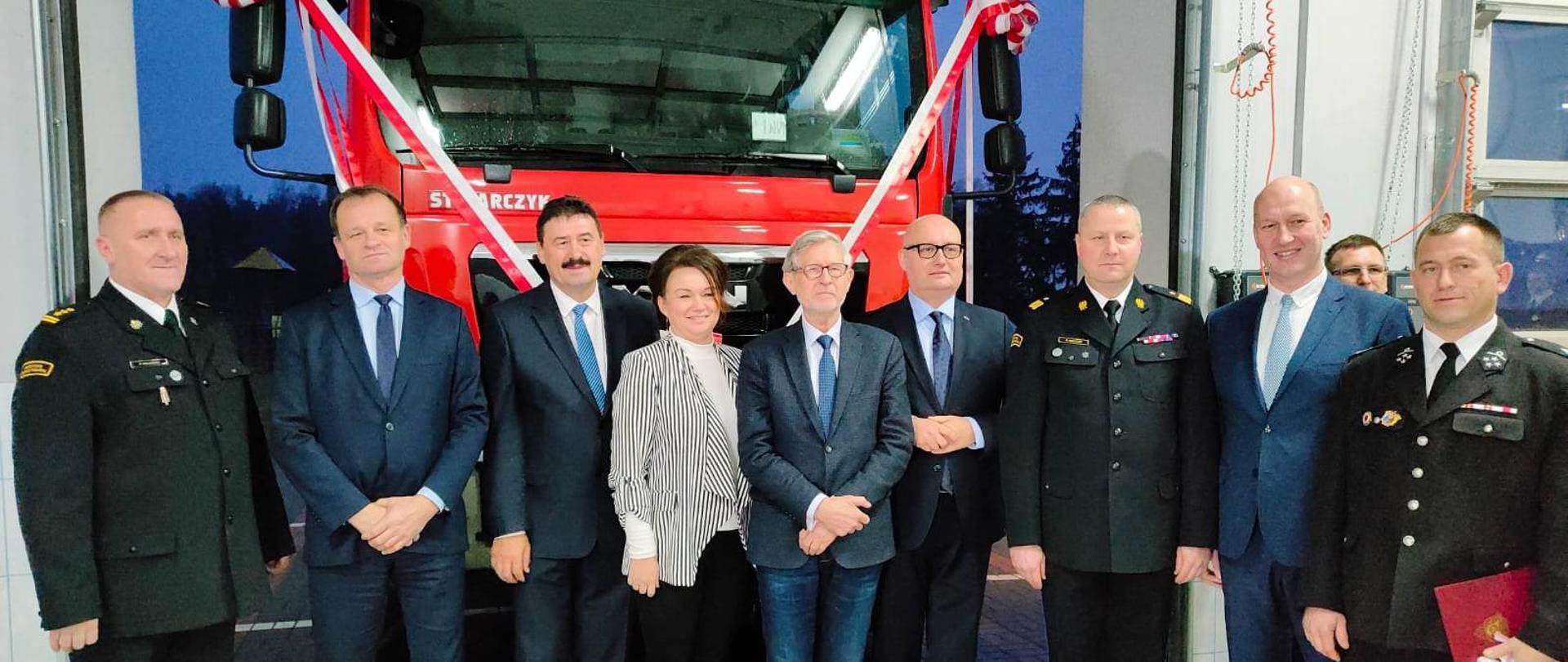 zdjęcie grupowe strażakó i osób cywilnych, w tle czedrwony samochó z biało czerwona wstega
