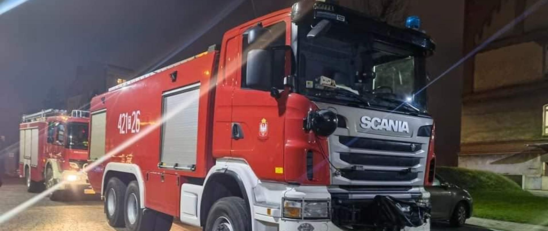 Pora nocna. Dwa czerwone wozy strażackie stojące na szarej kostce brukowej. W tle budynek mieszkalny.