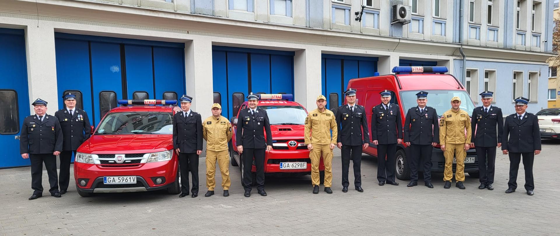 Strażacy PSP i OSP pozują do zdjęcia. Ubrani w mundury. W tle trzy czerwone samochody oraz budynek straży pożarnej.