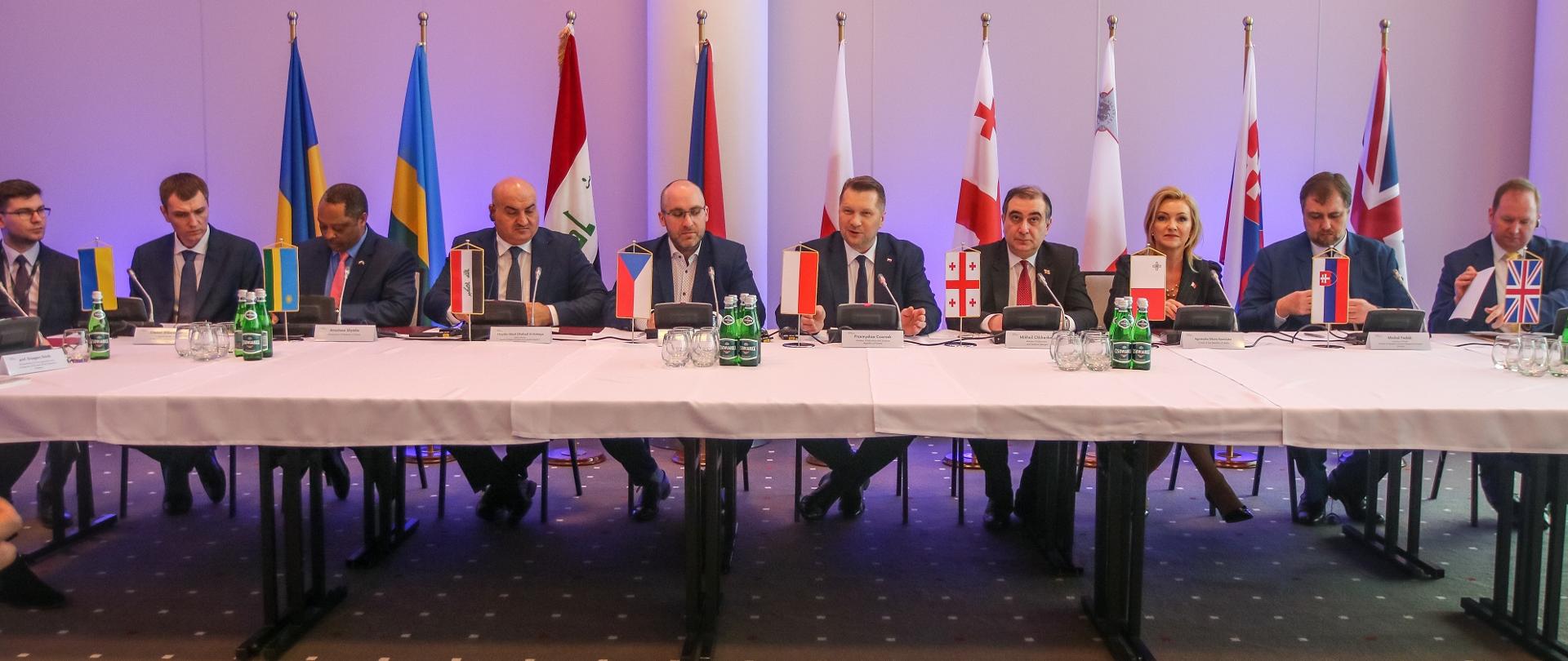 Przy szerokim nakrytym białą tkaniną stole siedzi kilkanaście osób, za nimi rząd flag różnych krajów i podświetlona na fioletowo ściana.