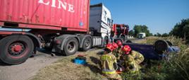 Na zdjęciu strażacy udzielają pomocy kierującej samochodem osobowym. Na drugim planie samochód osobowy koloru niebieskiego leżący w rowie na dachu. Po lewej stronie na jezdni stoi biały ciągnik siodłowy z przyczepą, na której załadowany jest czerwony kontener.