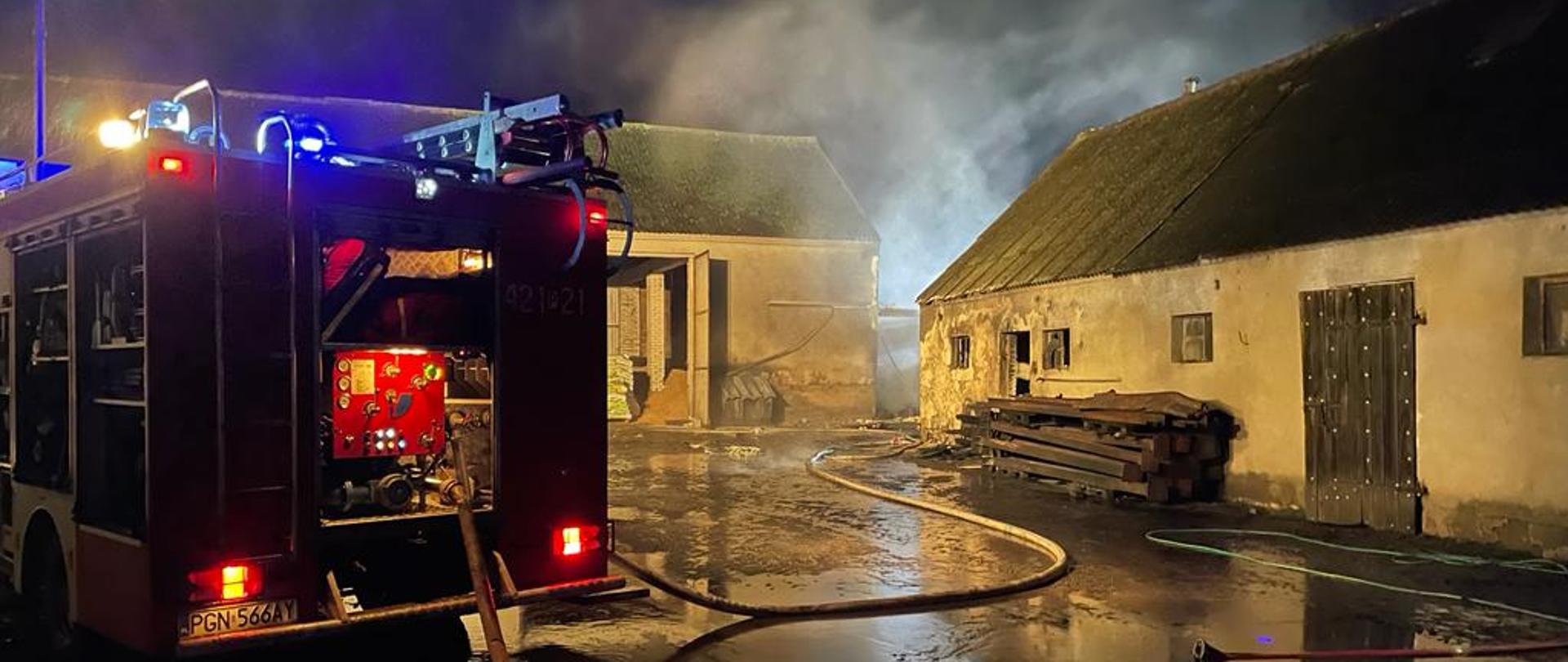 Na zdjęciu widać samochód pożarniczy. Jest pora nocna nad budynkami gospodarczymi unosi się dym