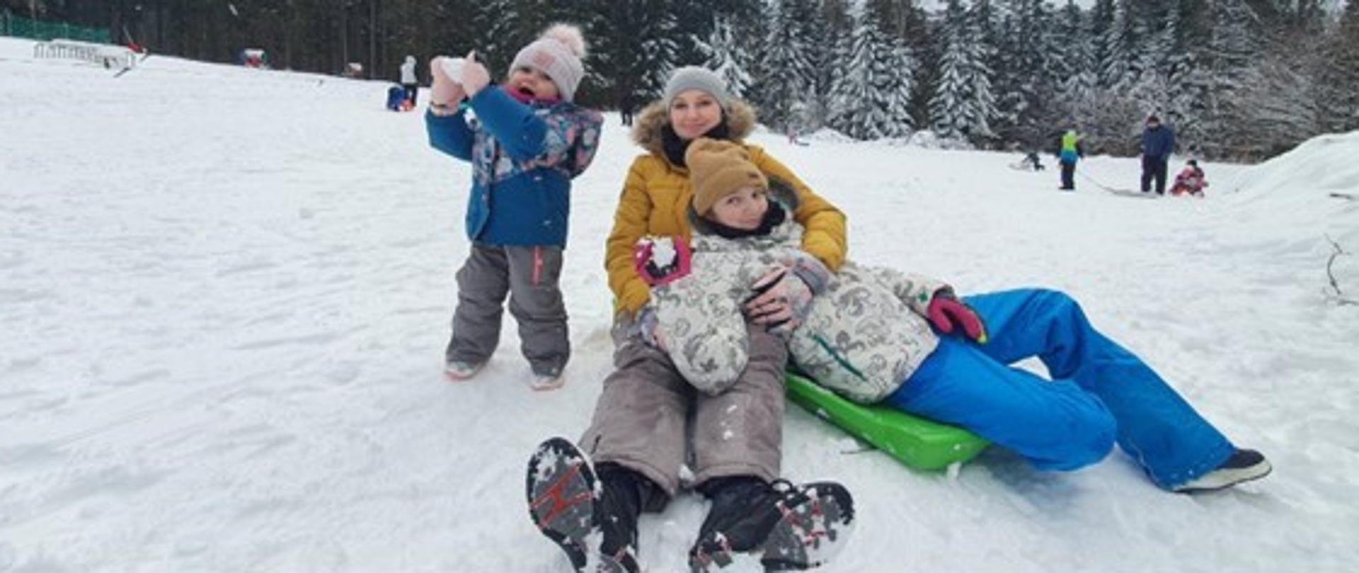 na pierwszym planie kobieta z dwójką dzieci na stoku śnieżnym. Po lewej stronie stoi dziewczynka , która trzyma kulkę śnieżną. Kobieta ze starszą dziewczynką siedzą na śniegu. Wszyscy są bardzo radośni. W tle las iglasty i mniej widoczne postaci osób stojących na stoku.
