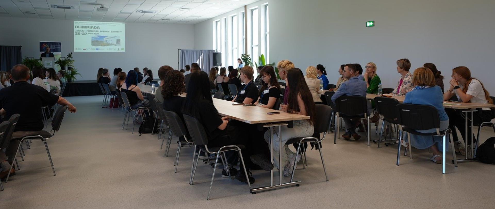 Duża sala konferencyjna z grupami osób siedzących przy stołach, skierowanych w stronę ekranu projektora. Na ekranie widoczny slajd z napisem ‘Olimpiada Biologia, Fizyka, Chemia i Ziemia 26–27 maja 2023’ oraz logotypami. W tle okna z naturalnym światłem