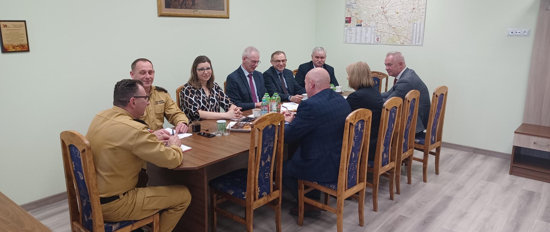 Na zdjęciu widać dwóch strażaków w żółtych strojach oraz dwie kobiety i pięciu mężczyzn. Wszyscy siedzą przy stole w pokoju. Na stole stoją napoje i ciastka. Na ścianie wisi obraz i mapa. Jest zapalone światło.