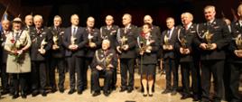 Grupa uhonorowanych strażaków i innych osób, statuetką z okazji Jubileuszu. Widok na kilkanaście osób w mundurach strażackich, harcerskim... Pośród stojących, na środku, na krześle siedzi druh senior.