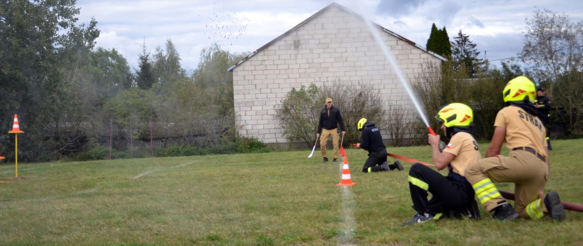 Na zdjęciu widać grupę młodych strażaków uczestniczących w ćwiczeniach na otwartej przestrzeni. Trzy osoby w hełmach strażackich, ubranych w beżowe koszulki z napisem „STRAŻ” oraz spodnie ochronne, obsługują wąż strażacki, z którego pod ciśnieniem leci strumień wody. Strumień skierowany jest na cel umieszczony na trawniku, zaznaczony pachołkami drogowo-ostrzegawczymi. W tle znajduje się mężczyzna obserwujący ćwiczenia, ubrany w czapkę z daszkiem i ciemną kurtkę. Całość odbywa się na trawiastym terenie w pobliżu jasnego budynku z białej cegły, otoczonego drzewami i krzewami. Niebo jest zachmurzone, co sugeruje pochmurny dzień.