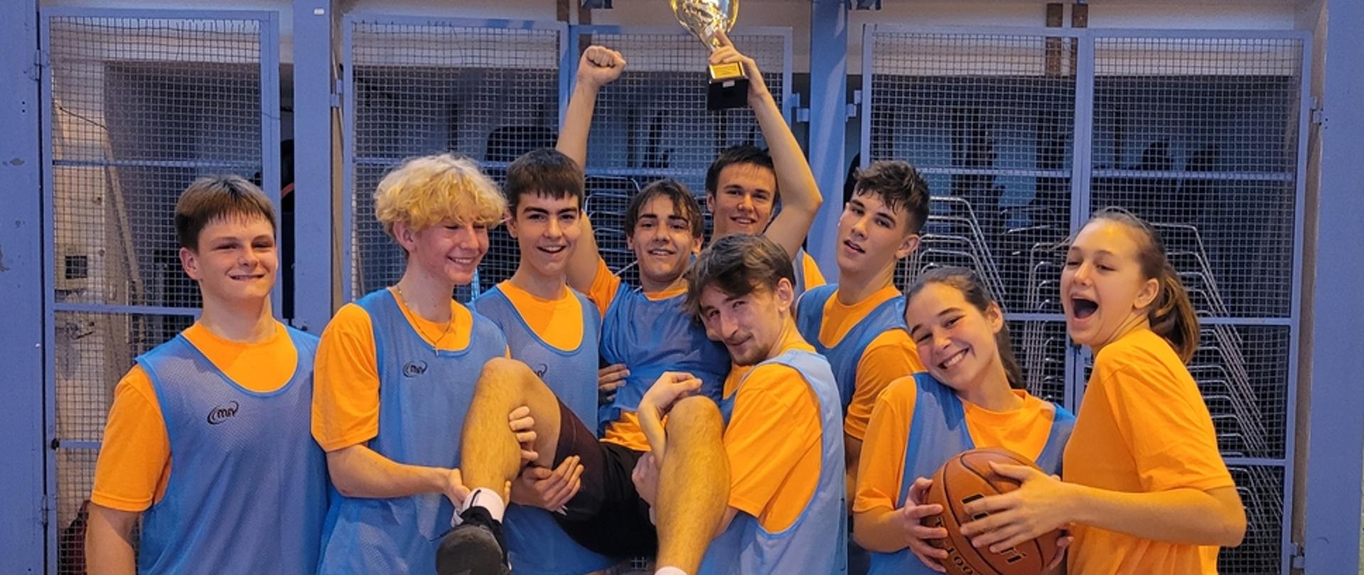 zdjęcie przedstawia drużynę w niebieskich koszulkach z pucharem zwycięstwa we wspólnym zdjęciu pod koszem na sali gimnastycznej