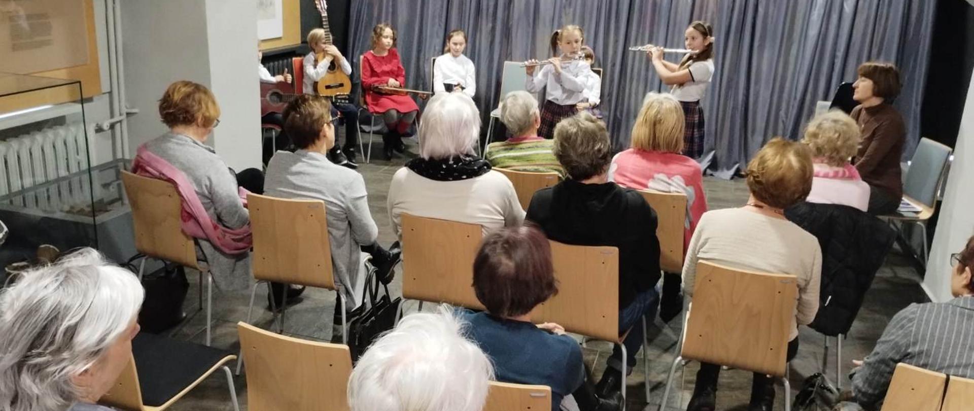 Zdjęcie z koncertu. Na scenie zaimprowizowanej scenie dzieci grające utwory. Siedząca na krzesłach publiczność to słuchacze Uniwersytetu Trzeciego Wieku. Z boku sceny opiekun uczniów, a także wychowawczyni zapowiadająca utwory. 