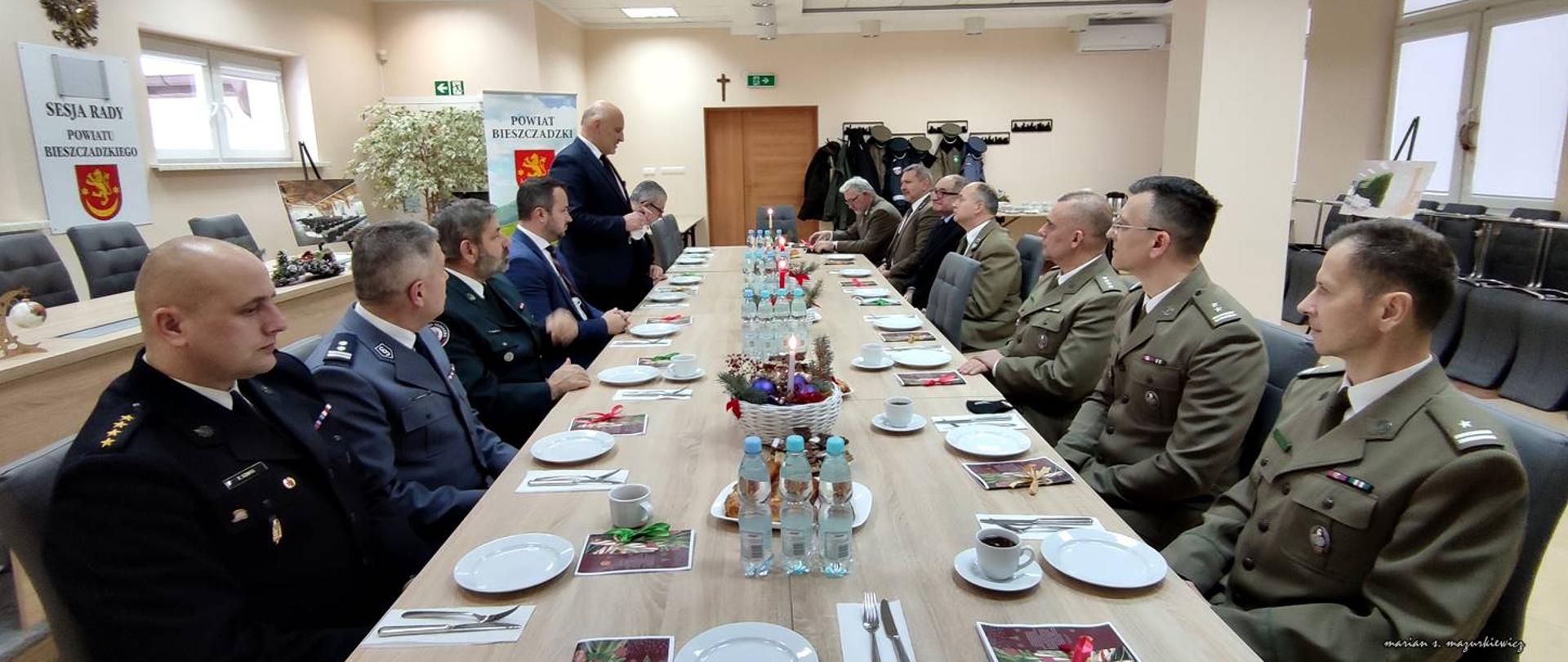 Na zdjęciu przedstawiciele służb mundurowych siedzący przy stole oraz Starosta Pan Marek Andruch - składający życzenia na stojąco. Zdjęcie wykonane jest w budynku starostwa, na stole znajdują się przekąski oraz woda w butelkach.