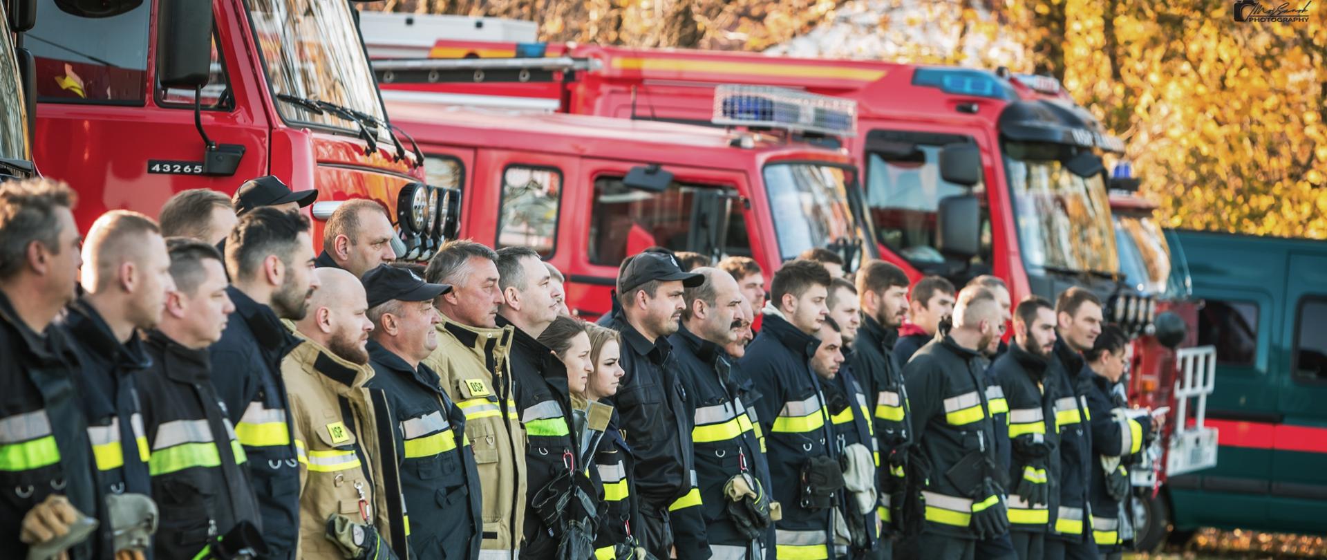 Zdjęcie przedstawia strażaków stojących na zbiórce przed ćwiczeniami. W tle samochody strażackie.