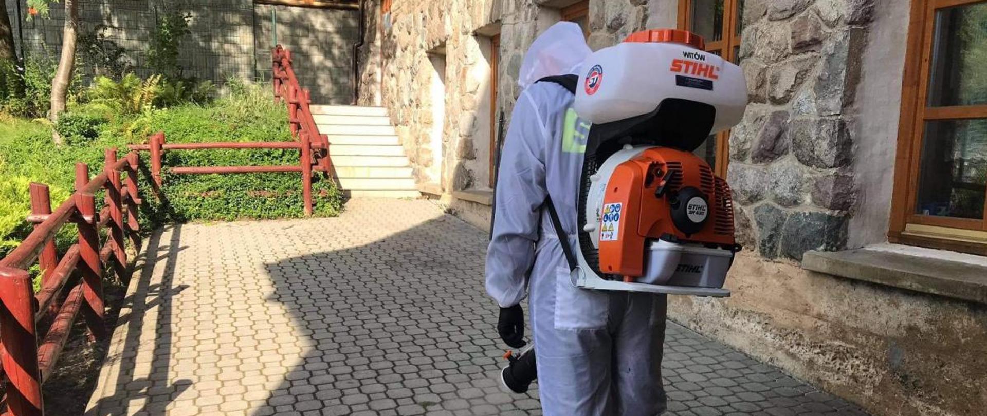 Na zdjęciu strażak stojący tyłem .Ubrany jest w biały kombinezon, na dłoniach ma czarne rękawiczki. Po jego prawej stronie znajduje się budynek wzdłuż którego umieszczone są okna, po lewej stronie znajduje się ogrodzenie- płot. Na wprost strażaka znajdują się schody przyległe do budynku.