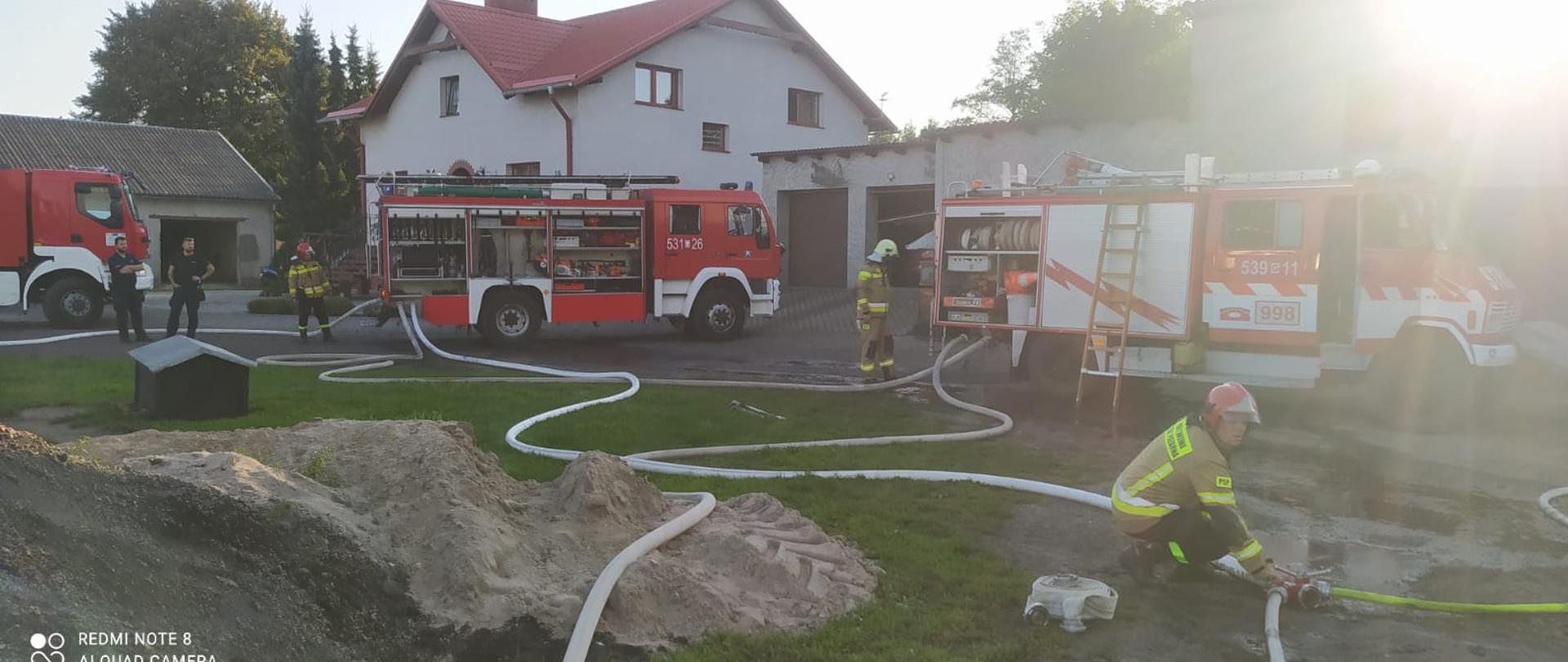 na placu widać 3 pojazdy pożarnicze z rozwiniętymi wężami i obsługujących sprzęt strażaków 