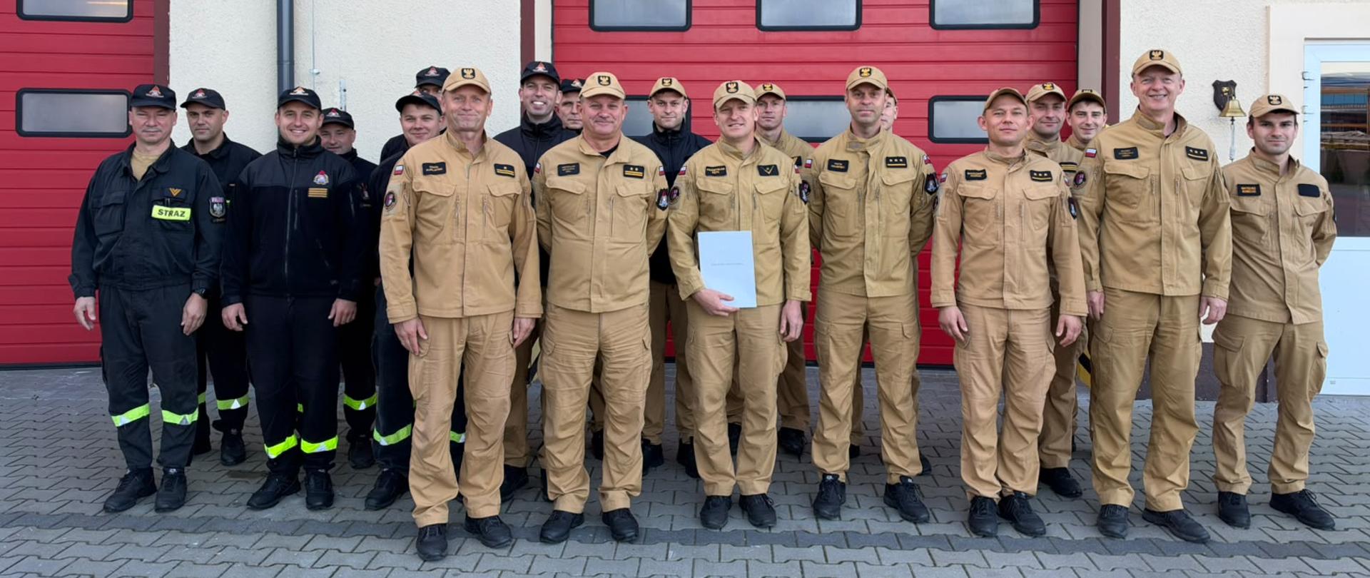 Zdjęcie przedstawia strażaków podczas awansu na stanowisko dowódcy sekcji. Strażace stoja na tle bram garazowych, ubrani sa w mundury piaskowe i czarne.
