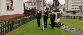Zdjęcie przedstawia trzech strażaków stojących przy maszcie podczas uroczystej zmiany służby, przed podniesieniem flagi państwowej. 