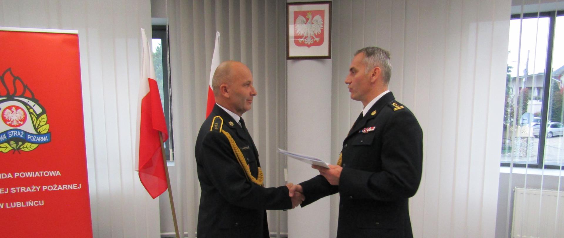 Zdjęcie zrobione w pomieszczeniu strażnicy na tle baneru Komendy Powiatowej PSP w Lublińcu oraz Flagi Państwowej. Na pierwszym planie dwóch strażaków z uściskiem dłoni w umundurowaniu galowym, zwróceni ku sobie.