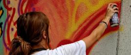 Stojąca tyłem dziewczyna z kitką, trzymająca w ręce spraj maluje po ścianie