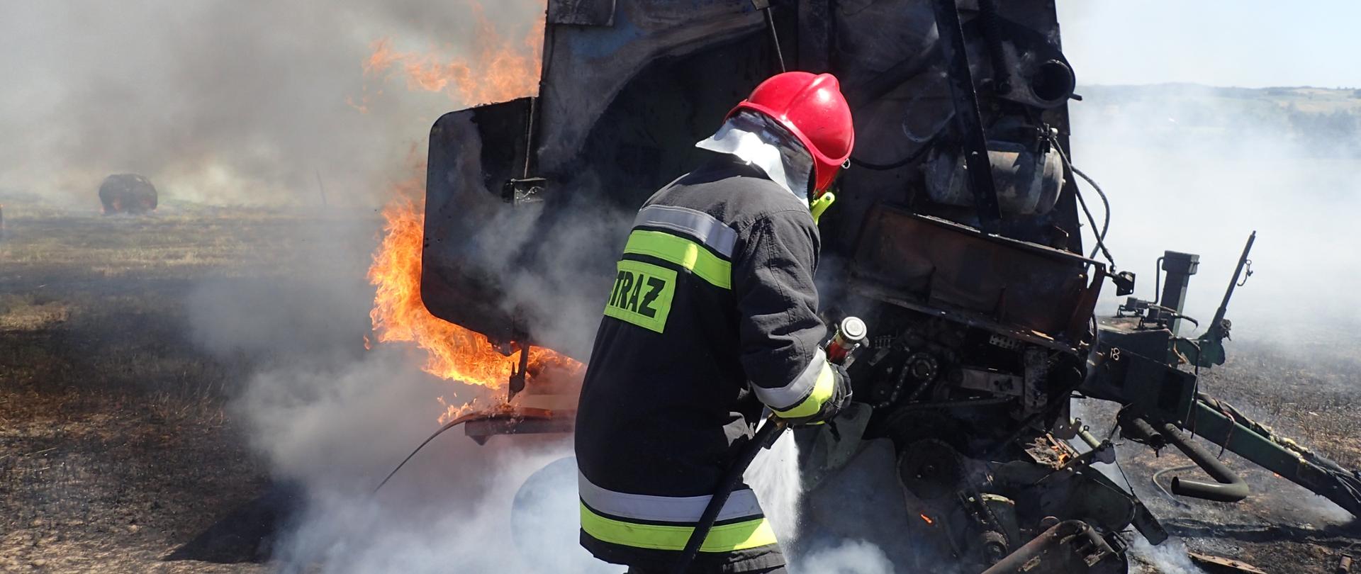 Zdjęcie przedstawia strażaka gaszącego pożar maszyny rolniczej podczas prac polowych