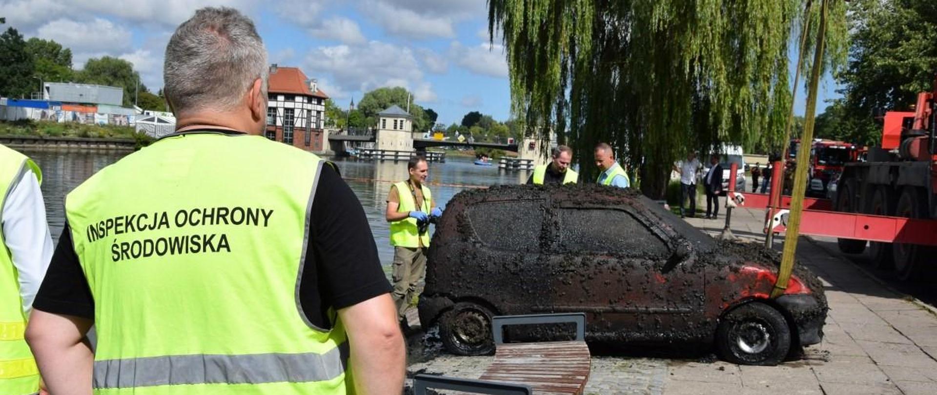 Na pierwszym planie Inspektor ochrony Środowiska w żółtej koszulce. Na chodniku stoi ubłocony, czerwony Fiat seicento, któremu przyglądają się funkcjonariusze. W tle zielona wierzba płacząca i most.