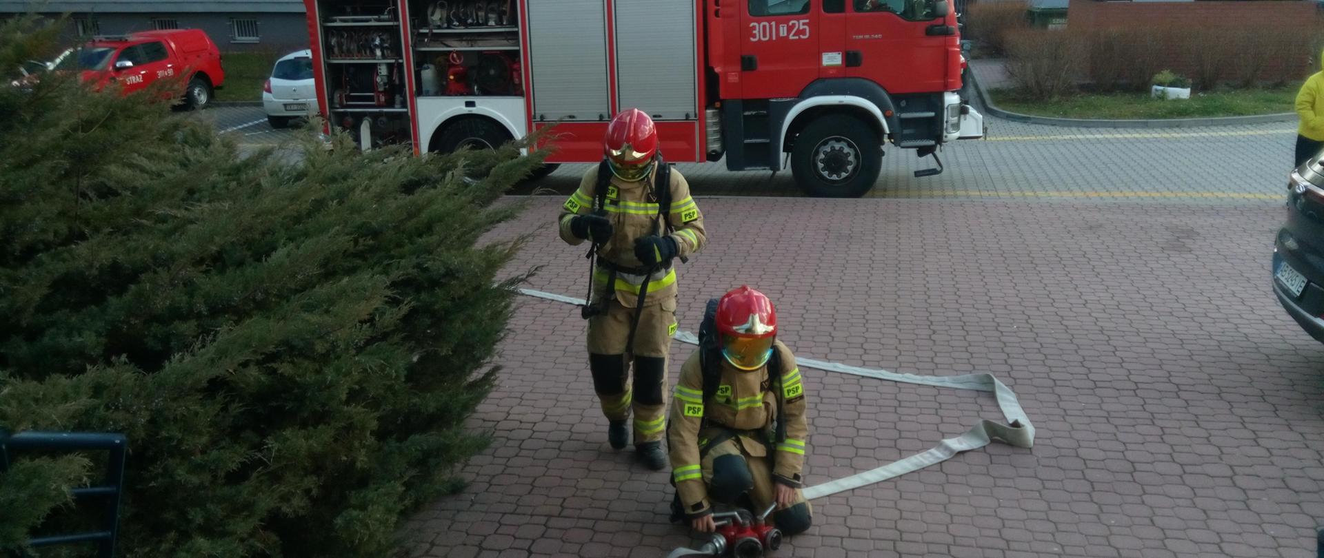 Zdjęcie przedstawia strażaka przy rozdzielaczu. Za nim ratownik ubiera się w sprzęt ochrony układu oddechowego. Za nimi znajduje się samochód strażacki.