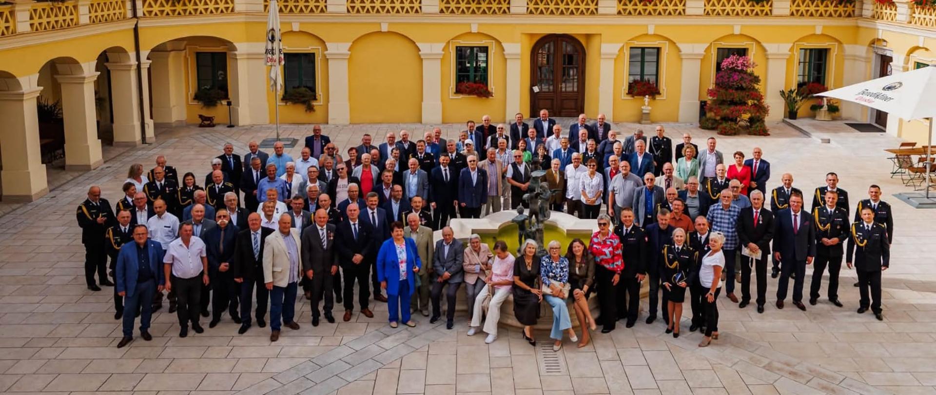 Duża grupa uczestników Jubileuszu stoi i siedzi wokół ozdobnej fontanny na dziedzińcu Pałacu w Żyrowej. Budynek w tle ma żółte elewacje, zielone okiennice i arkadowe podcienia. Widać osoby w garniturach, mundurach PSP oraz członków ZEiRP RP. Uczestnicy pozują do wspólnego zdjęcia, a przestrzeń jest jasna, zadbana i udekorowana kwiatami.