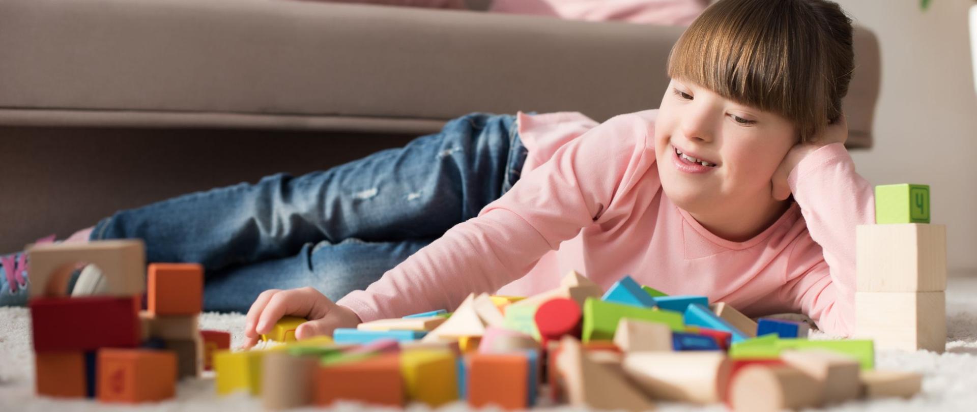 Na zdjęciu jest dziewczynka leżąca na podłodze i układająca klocki.