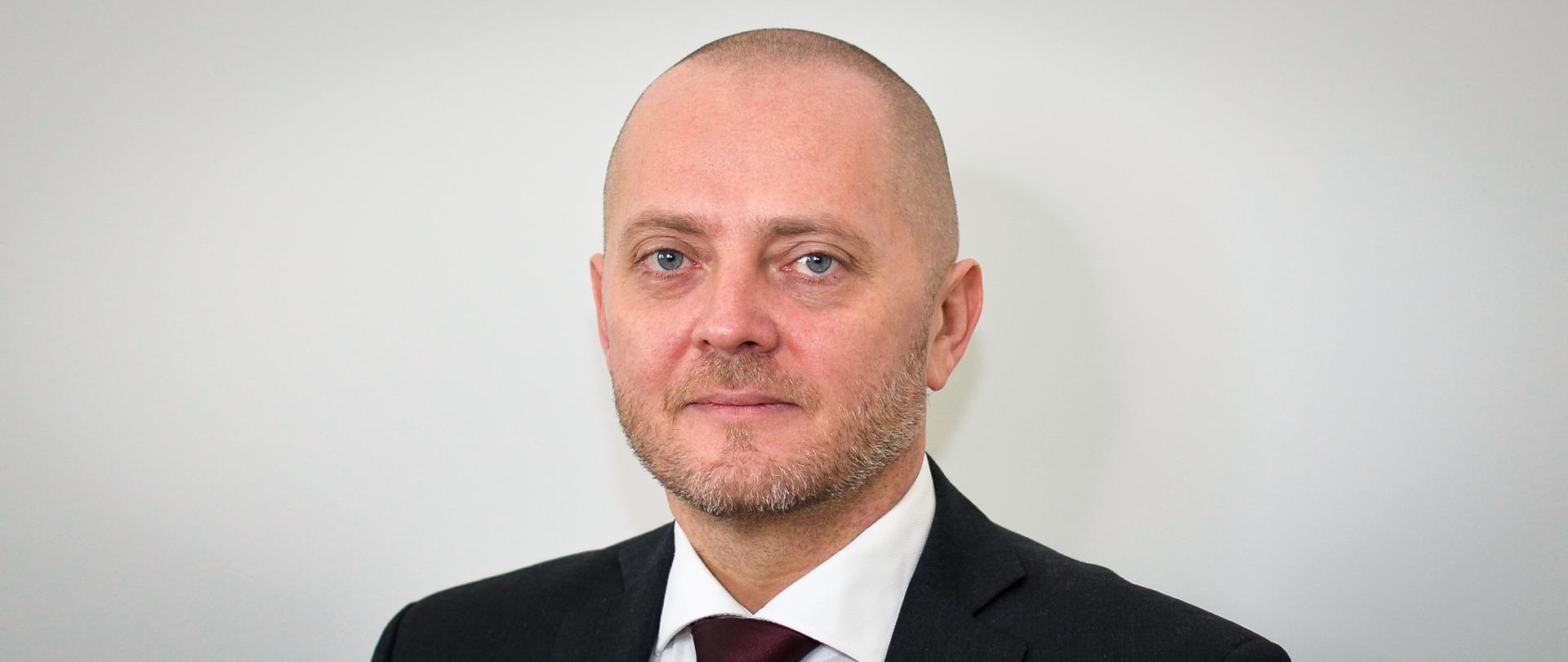 Mężczyzna w ciemnej marynarce, białej koszuli, bordowym krawacie, z krótkim zarostem.