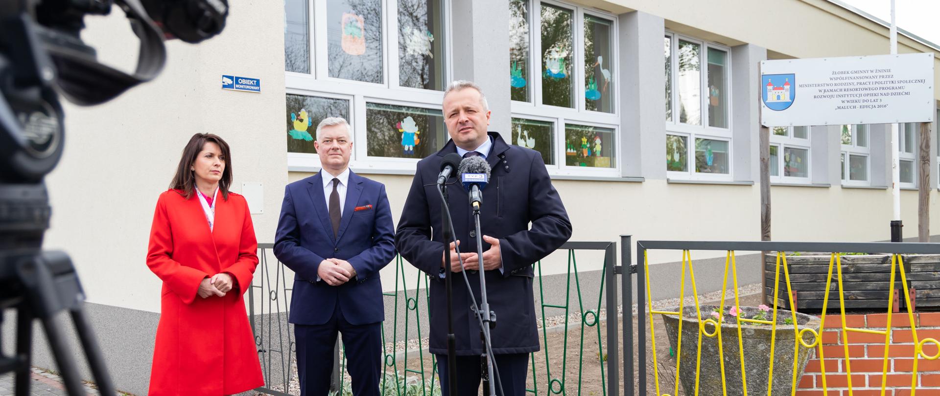 Jeden mężczyzna stojący przy mikrofonie i wypowiadający się. Za nim kobieta i mężczyzna