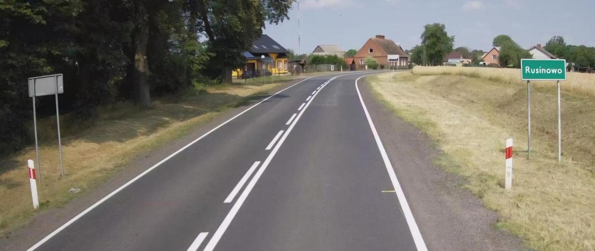 Na zdjęciu widoczna jest asfaltowa droga prowadząca przez wieś. Droga ma wyraźnie oznaczone białe linie, w tym przerywaną linię środkową i ciągłe linie boczne. Po lewej stronie drogi znajduje się znak drogowy z napisem Rusinowo wskazujący nazwę miejscowości. W tle widać kilka domów otoczonych drzewami i polami, co sugeruje wiejski krajobraz.