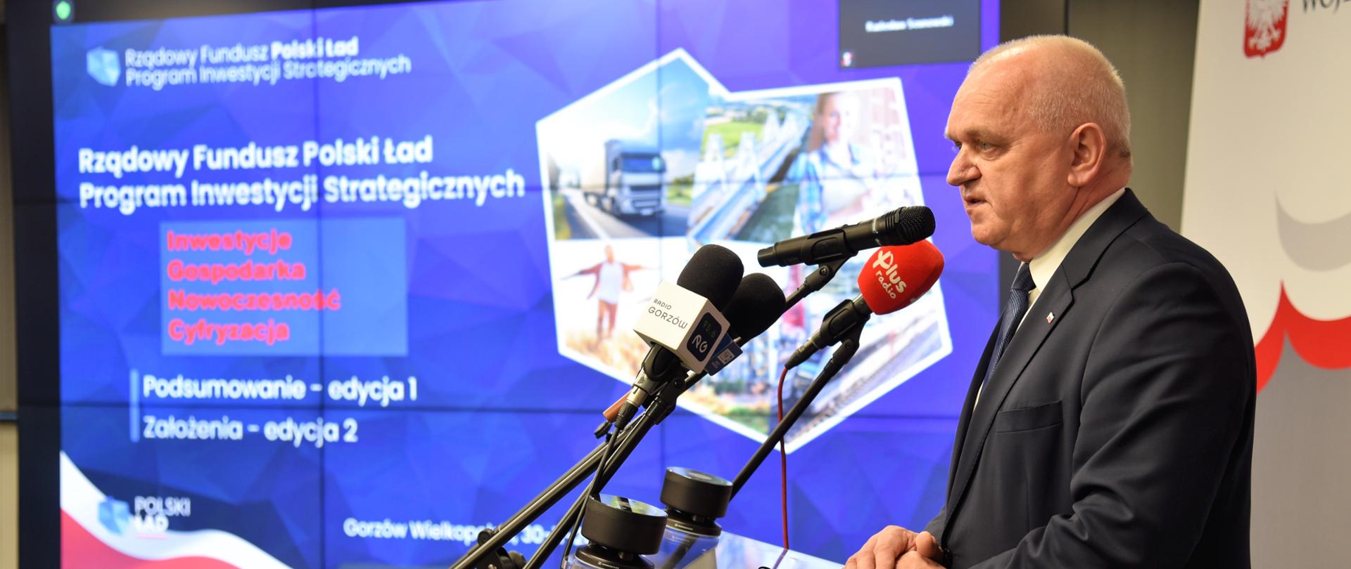 Wojewoda stoi przy mównicy - widok z profilu. W tle slajd prezentacji 